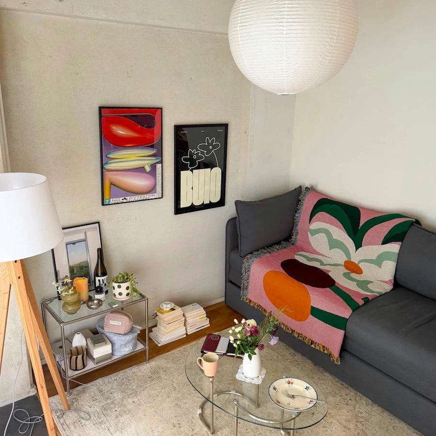 A cozy living room corner featuring a gray sofa with a colorful pink, green, and orange abstract floral throw blanket, a glass coffee table, a white paper lantern pendant light, and framed abstract wall art.