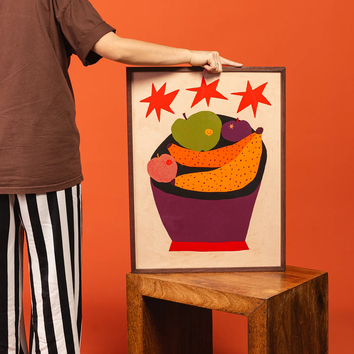 A framed art print featuring a colorful fruit bowl illustration with red stars, held by a person wearing a brown shirt and black-and-white striped pants against a solid orange background.