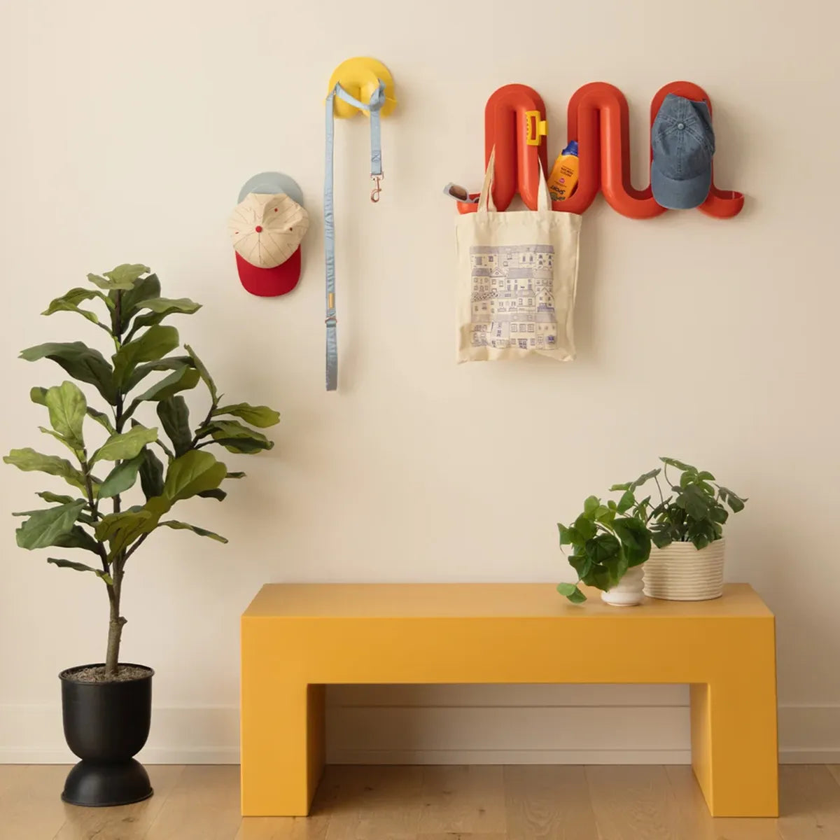 A styled entryway scene with a mustard yellow bench against a cream wall, a red Heller Swell Wall Catchall holding hats, a leash, and a tote bag, plus potted plants on the floor.