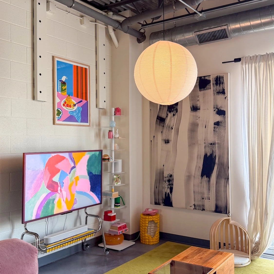 A modern living room interior with colorful abstract wall art, a large black-and-white vertical canvas, a TV on a metal stand, a white paper lantern pendant light, shelving, curtains, and a wooden coffee table.