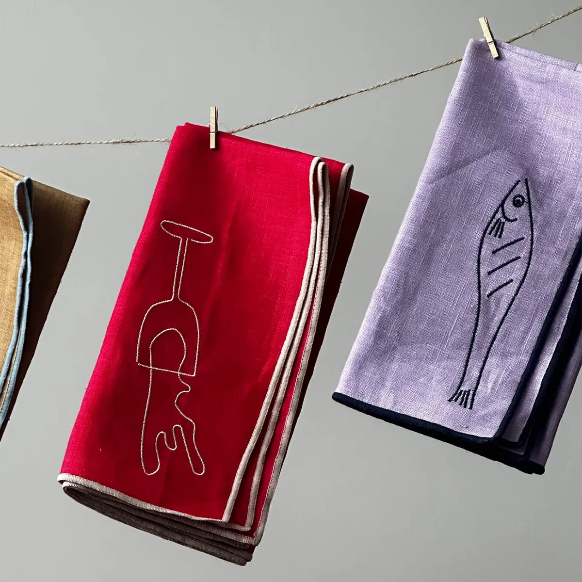 Red napkin with wine glass design and purple napkin with fish design hanging on a line against a gray background.