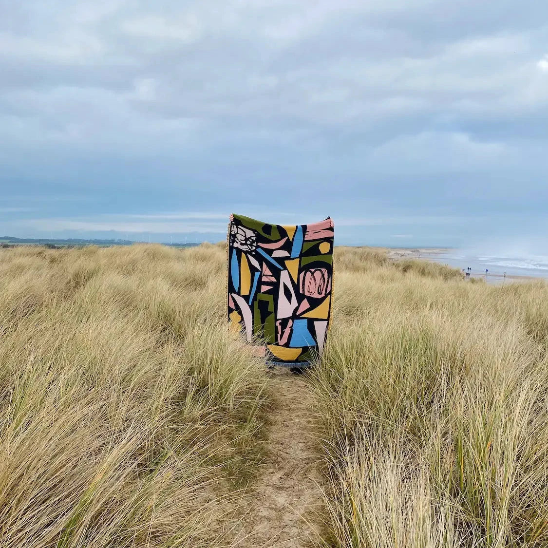 Colorful patterned blanket draped over a grassy dune with ocean and sky in the background