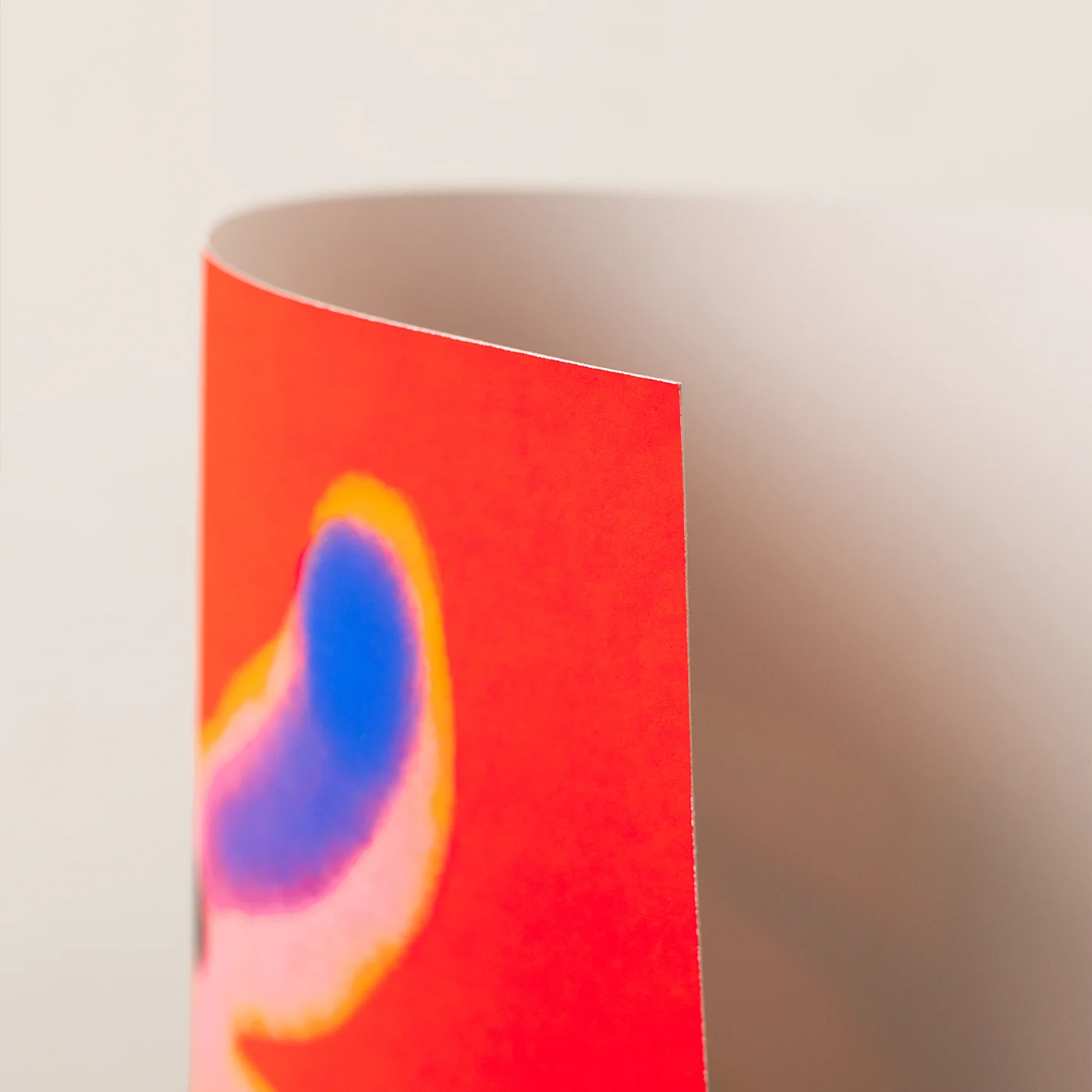 A close-up of an art print corner showing a bright red surface with an abstract blue, purple, and yellow shape, photographed against a soft beige background.