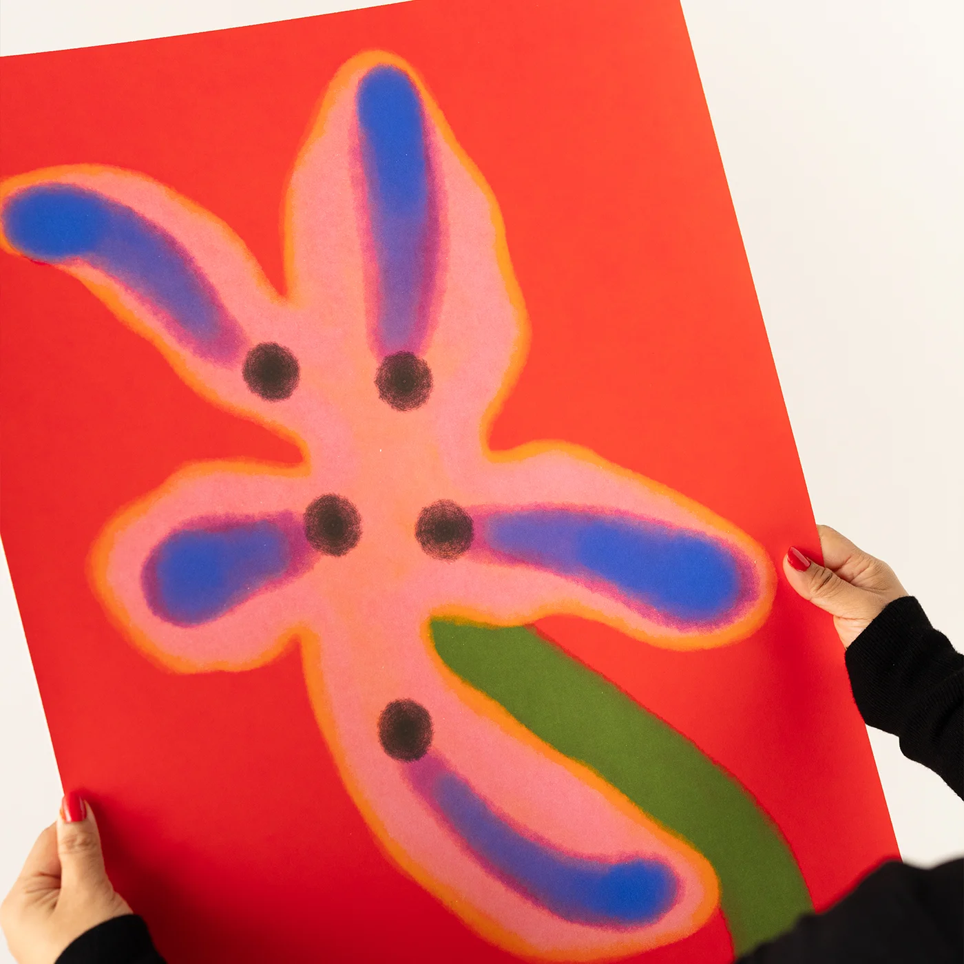 A person holds a vibrant red art print featuring a stylized flower with pink petals, blue accents, and a green stem. The hands holding the print are visible at the corners against a clean white background.