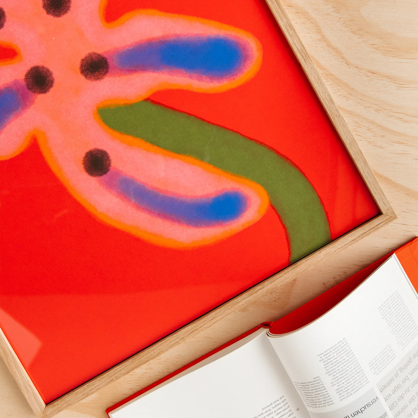 A framed abstract art print featuring a bold red background with a pink flower-like shape outlined in orange, blue accents, and a green stem, displayed on a light wood surface next to an open book.