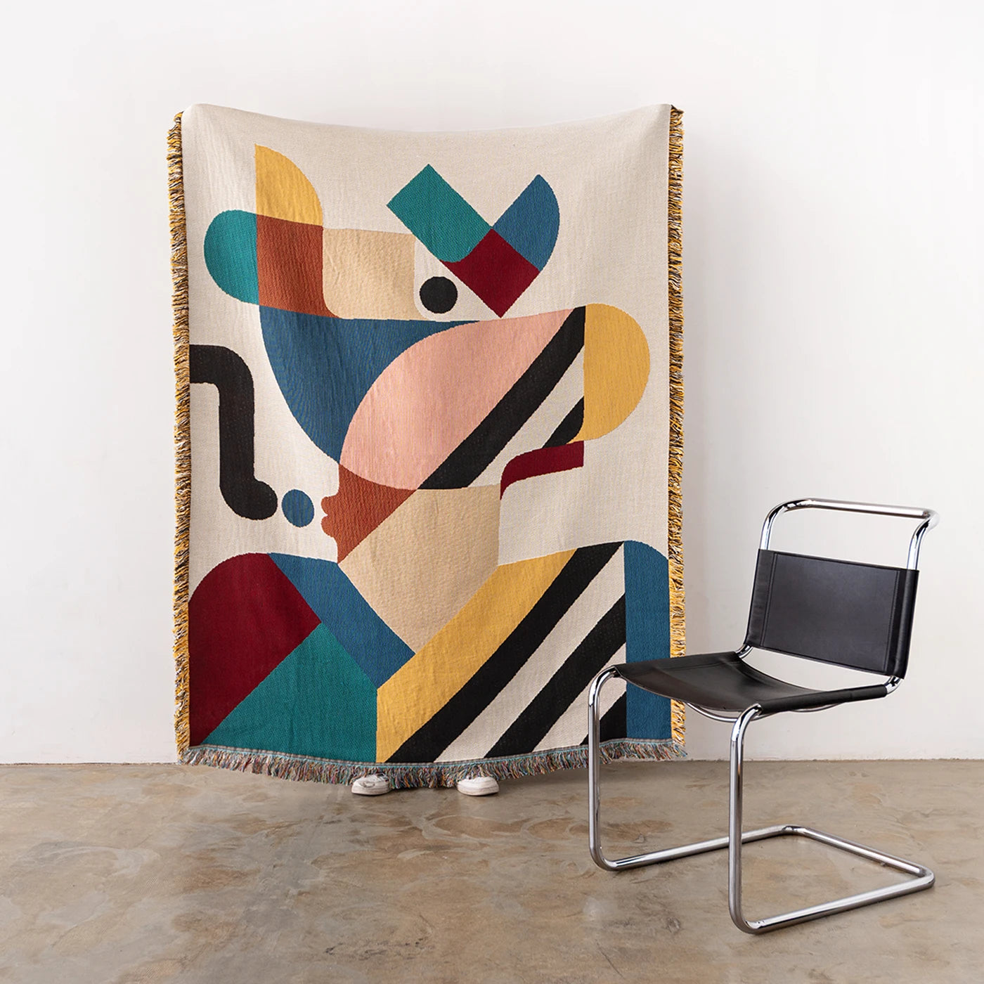 A large woven throw with an abstract geometric colourful design hanging against a white wall in a room. A black chair with a chrome frame sits to the right.