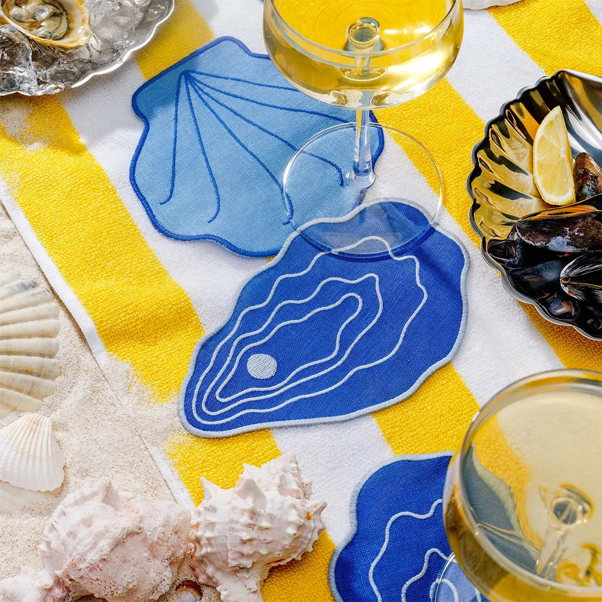Table setting with blue shell coasters, white wine glasses, and seashells on a yellow and white tablecloth.
