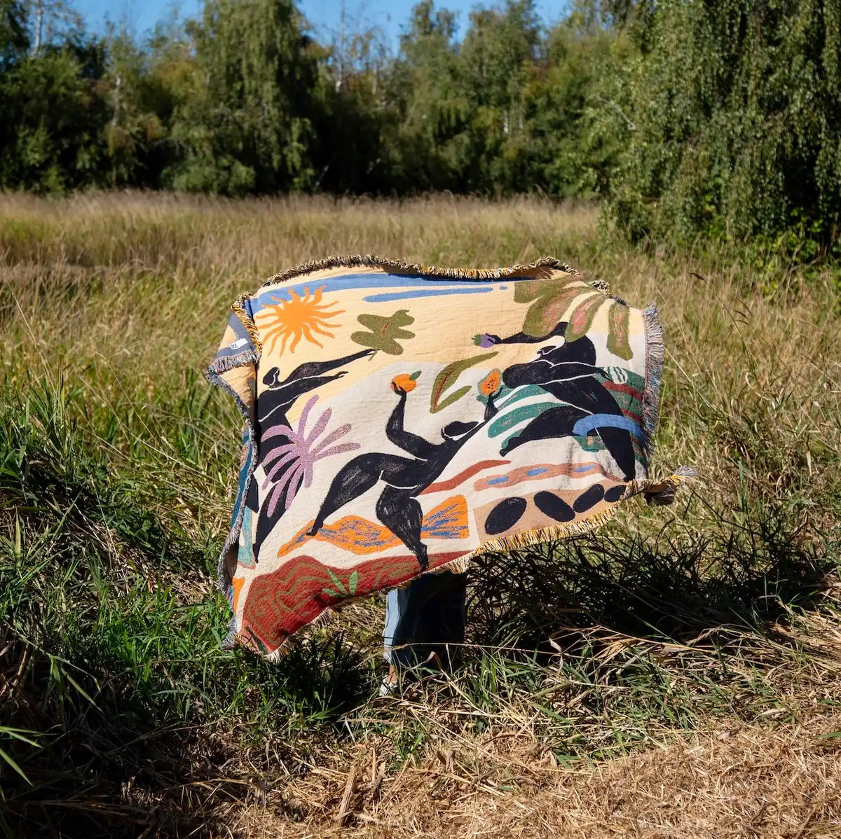 A large woven throw blanket with fringe is held up against a natural landscape with tall grass. The blanket features a colorful, abstract folk-art style design with black human figures, a yellow sun, and green and blue botanical elements on a cream base.