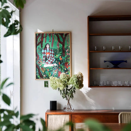 A colorful framed art print depicting a seated figure in a forest, displayed on a white wall above a wooden sideboard in a modern interior setting with plants and shelves.