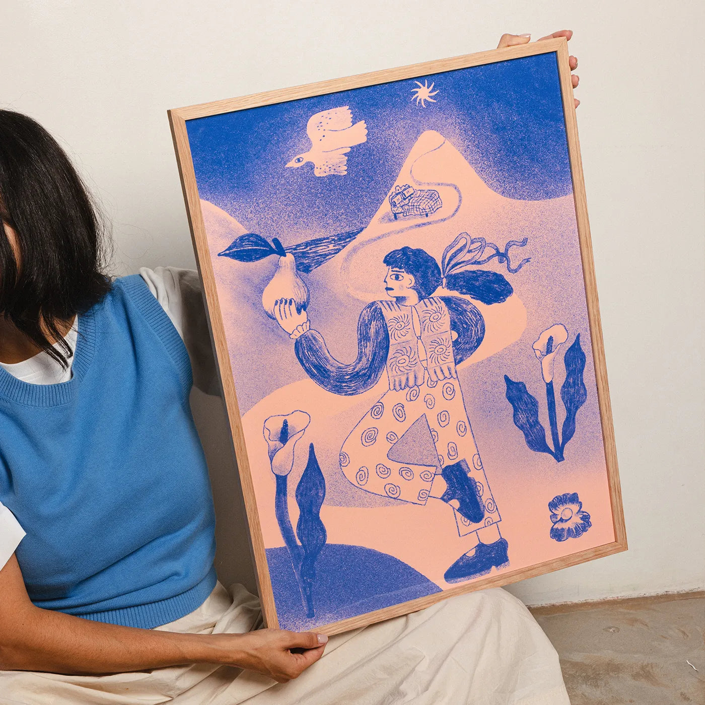 A person seated indoors holds a large framed art print. The artwork features a stylized figure in blue against a soft pink-to-blue gradient background with abstract floral and bird motifs.