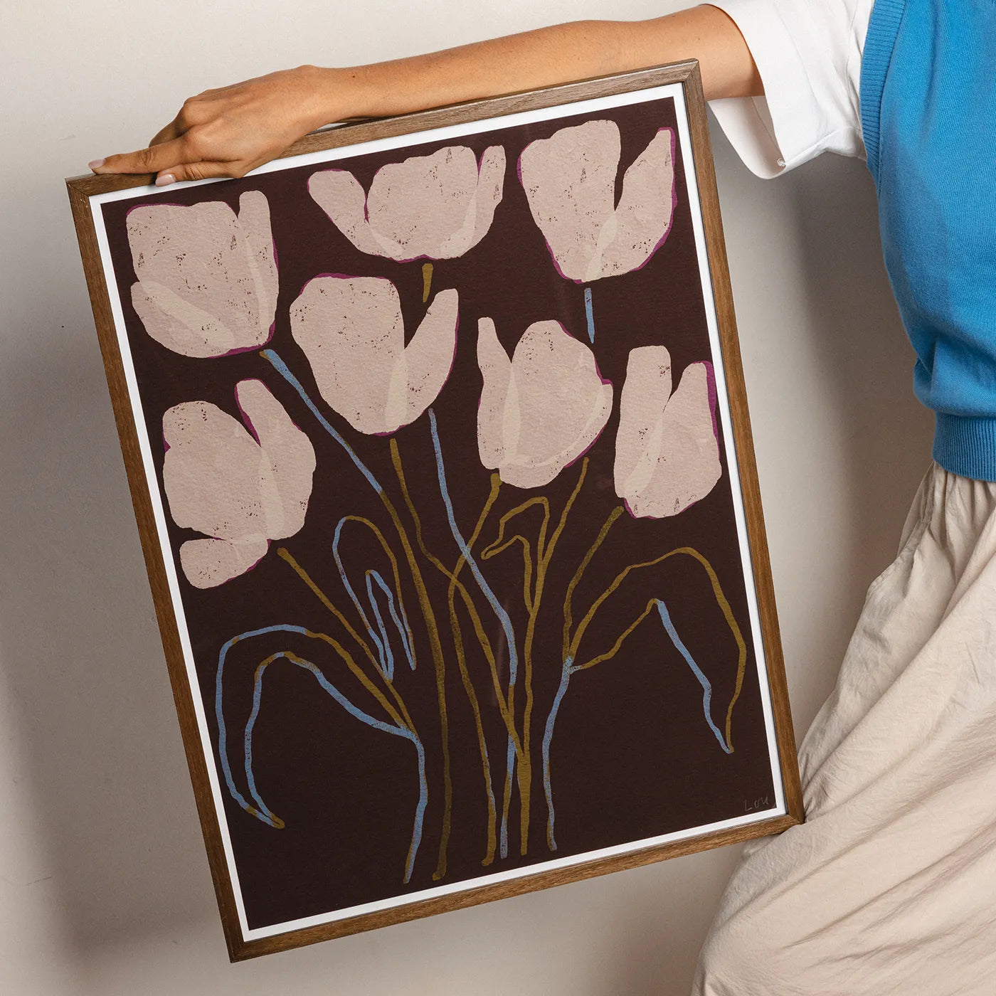A framed floral art print featuring pale pink tulip-like flowers on a dark brown background, held by a person wearing a blue top and light pants against a plain light gray wall.