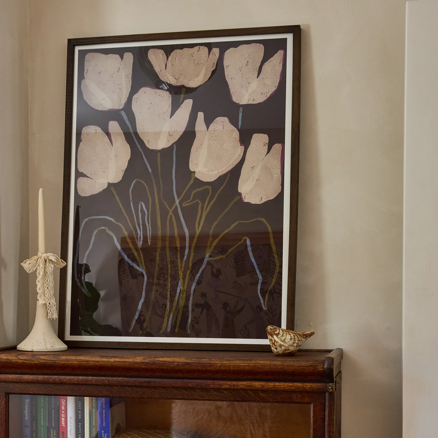 A framed floral wall art piece with large beige tulip-like shapes on a dark background, displayed leaning against a beige wall above a wooden cabinet, with a candle and small decorative object nearby.