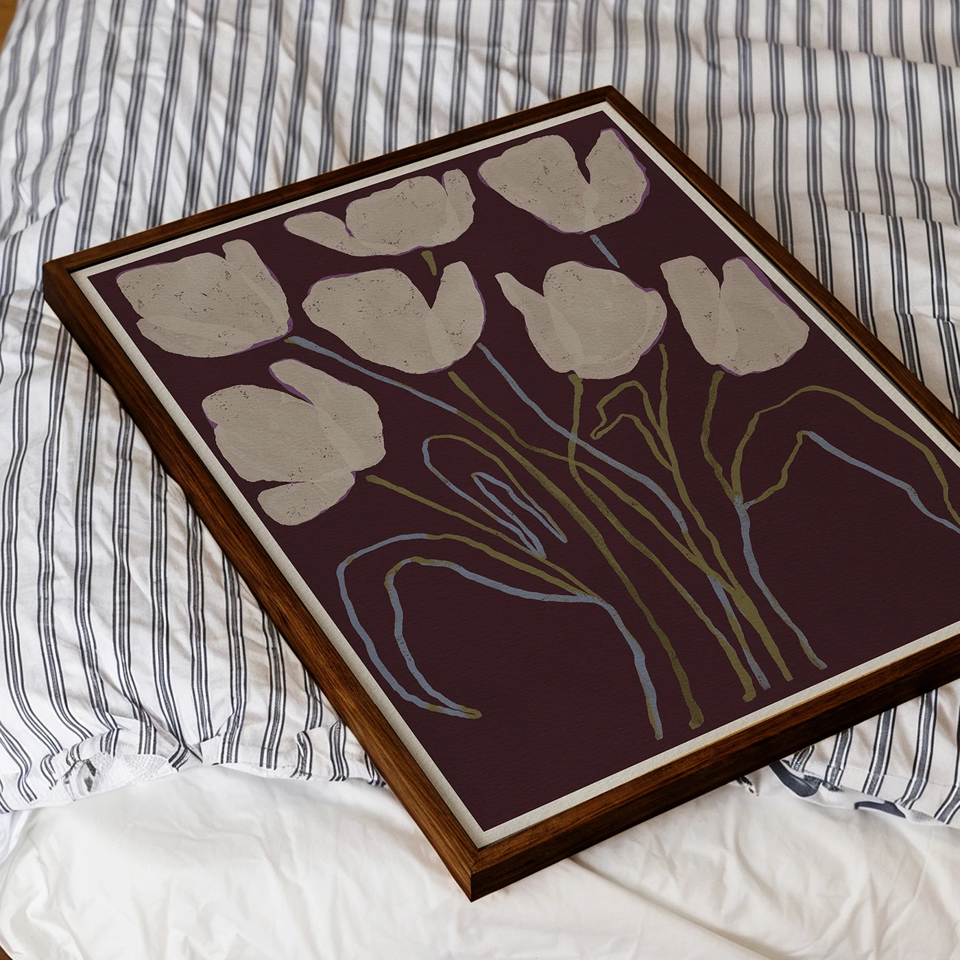 A framed floral art print featuring stylized tulips in cream and light blue on a dark plum background, resting on a bed with white and navy striped bedding.