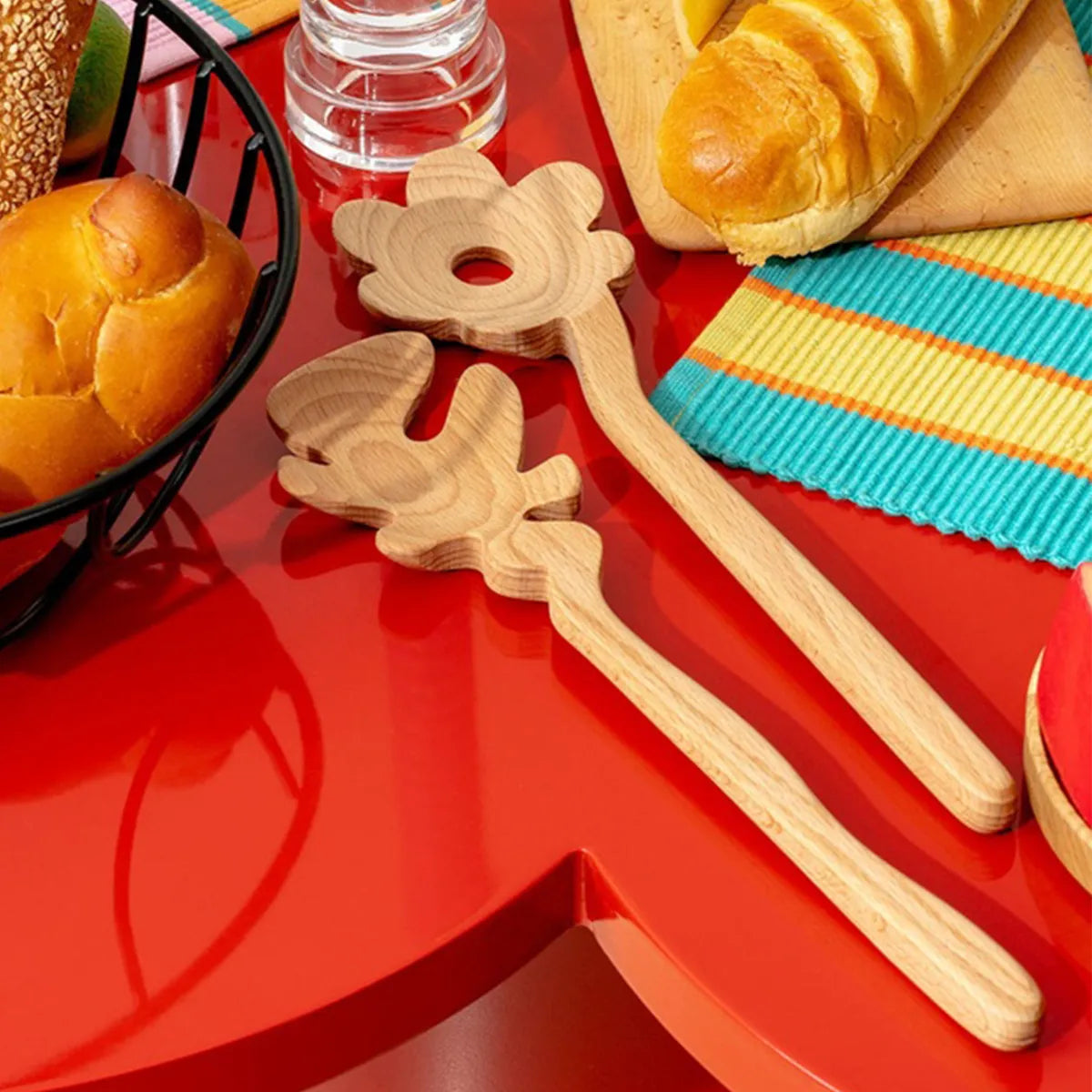 Two light natural wood flower-shaped Serving Friends spoons by Areaware, displayed on a glossy red tray with bread and a striped napkin in the background.