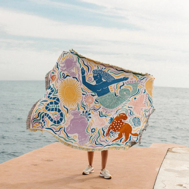 Person holding a colorful bed blanket with ocean and sun design on a dock.