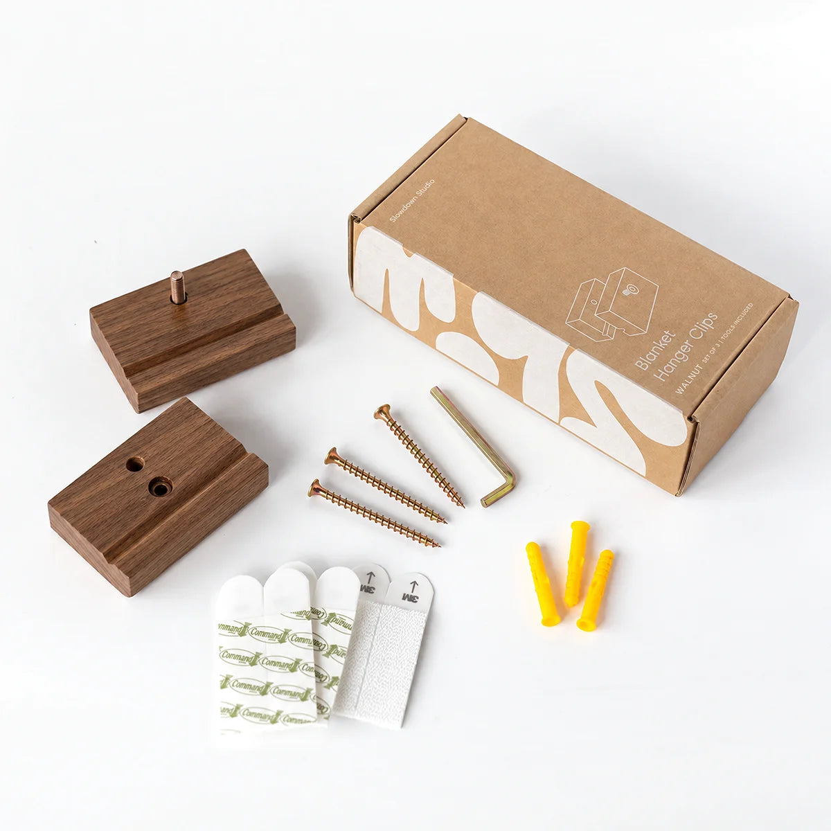 A blanket hanger set of 3 clips in walnut with a  kit laid out on a white background, including 3 screws, 3 wall anchors, 3 white adhesive hooks, an Allen key and a brown packaging box.