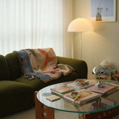 Living room with a green sofa, orange and green blanket for couch and glass coffee table, and framed picture on the wall.