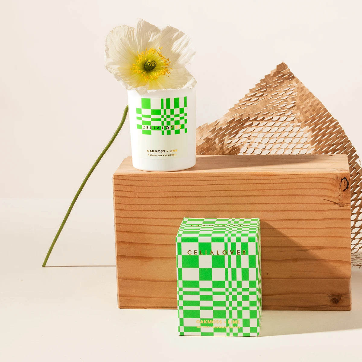 Candle with green and white checkered design on a wooden block with a flower and packaging box.