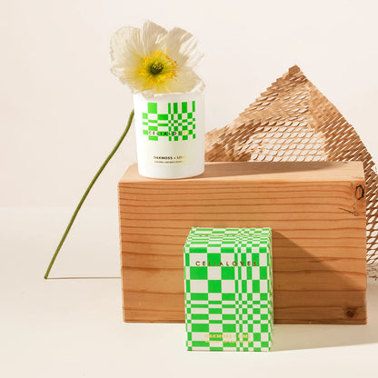 Candle with green and white checkered design on a wooden block with a flower and packaging box.