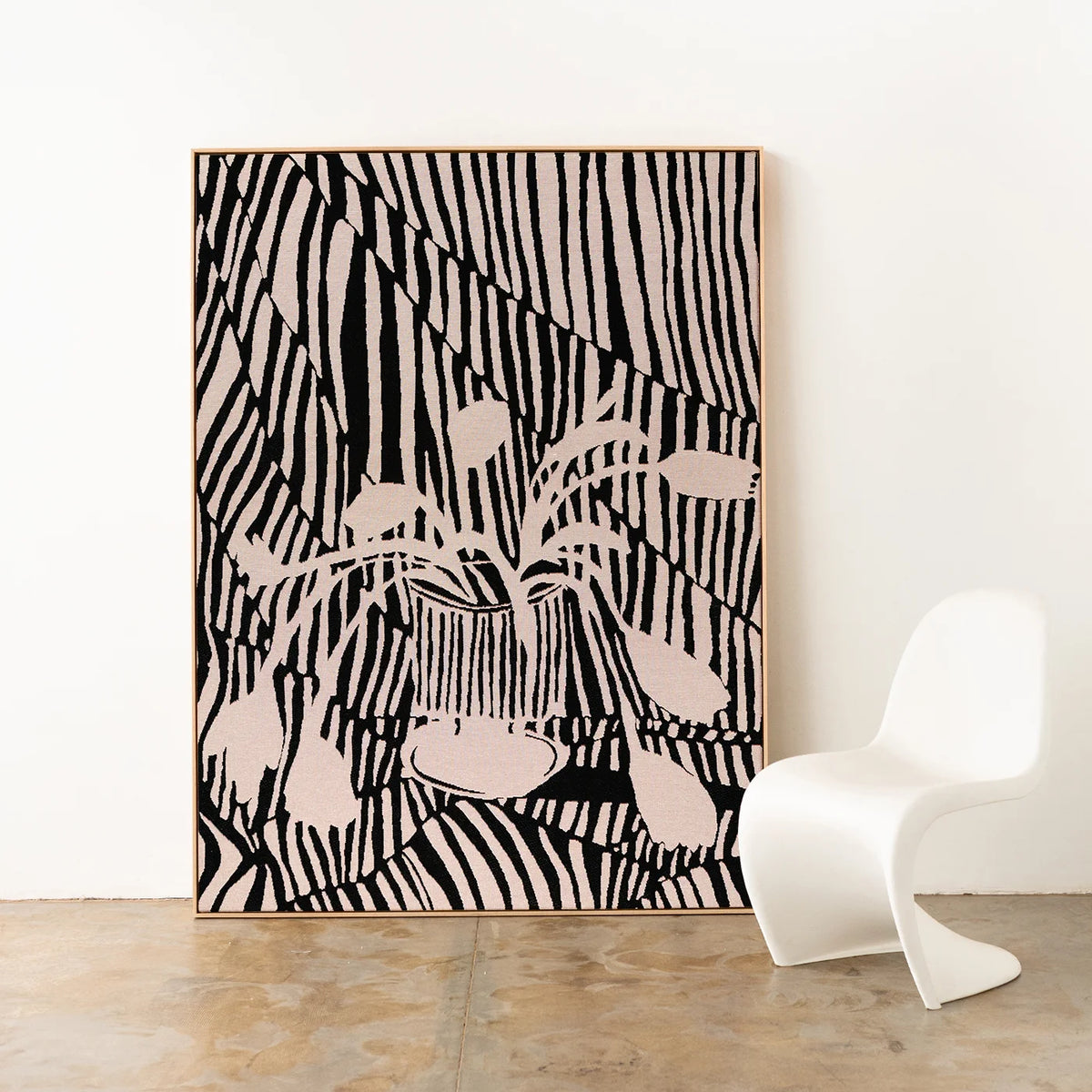 Framed large wall art featuring a black and beige zebra-like pattern and a central floral motif, leaning against a white wall on a concrete floor next to a white molded chair.