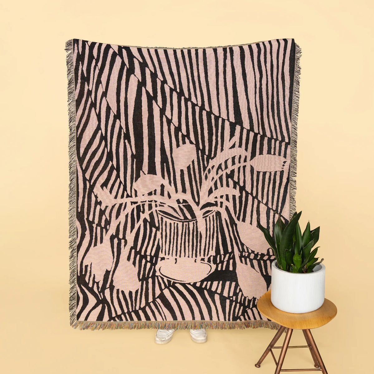 A woven throw blanket with a pink and black zebra-stripe pattern and a central floral motif, styled on a beige background next to a small potted plant on a wooden stool.