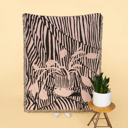 A woven throw blanket with a pink and black zebra-stripe pattern and a central floral motif, styled on a beige background next to a small potted plant on a wooden stool.