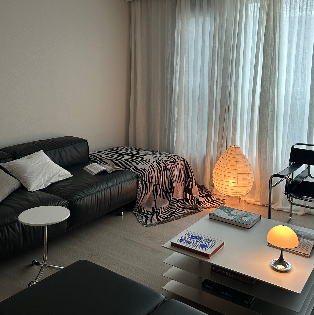 A modern living room corner featuring a black leather sofa with white pillows, a black and white striped throw blanket sofa cover, a white side table, a white coffee table with books, and a glowing paper lantern lamp near sheer white curtains.
