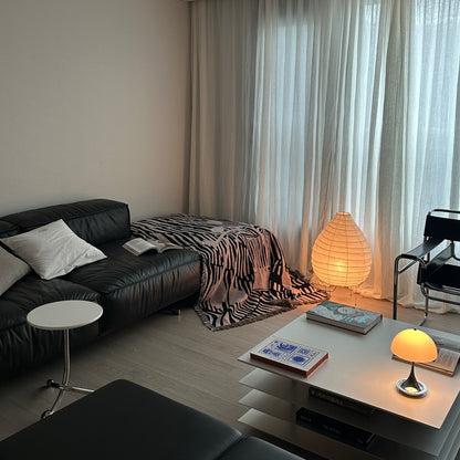 A modern living room corner featuring a black leather sofa with white pillows, a black and white striped throw blanket sofa cover, a white side table, a white coffee table with books, and a glowing paper lantern lamp near sheer white curtains.