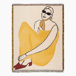 Slowdown Studio woven throw blanket with fringe on all sides, featuring a minimalist line-art illustration of a seated woman in a yellow dress, orange scarf, black sunglasses, and red shoes on a cream background.