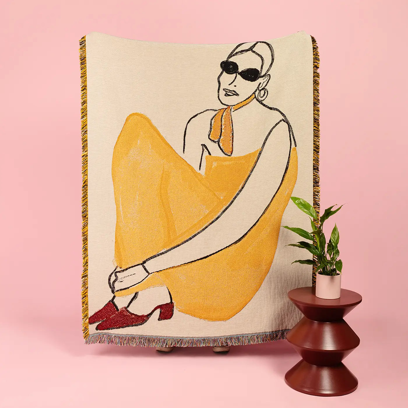 A woven throw blanket featuring a stylized illustration of a woman in a yellow dress and red shoes, displayed on a pink background next to a small potted plant on a wooden stand.
