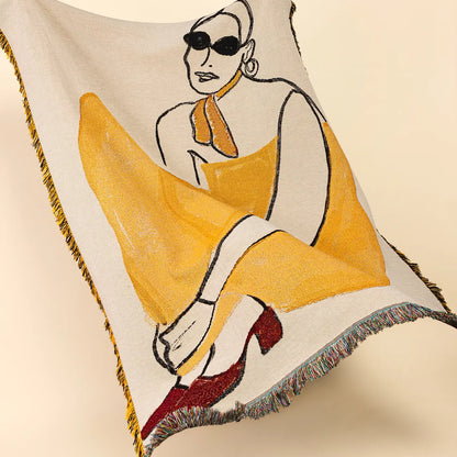 A cream-colored woven throw blanket with fringe edges, featuring a stylized illustration of a woman in a yellow dress and red heels, displayed against a solid beige background.