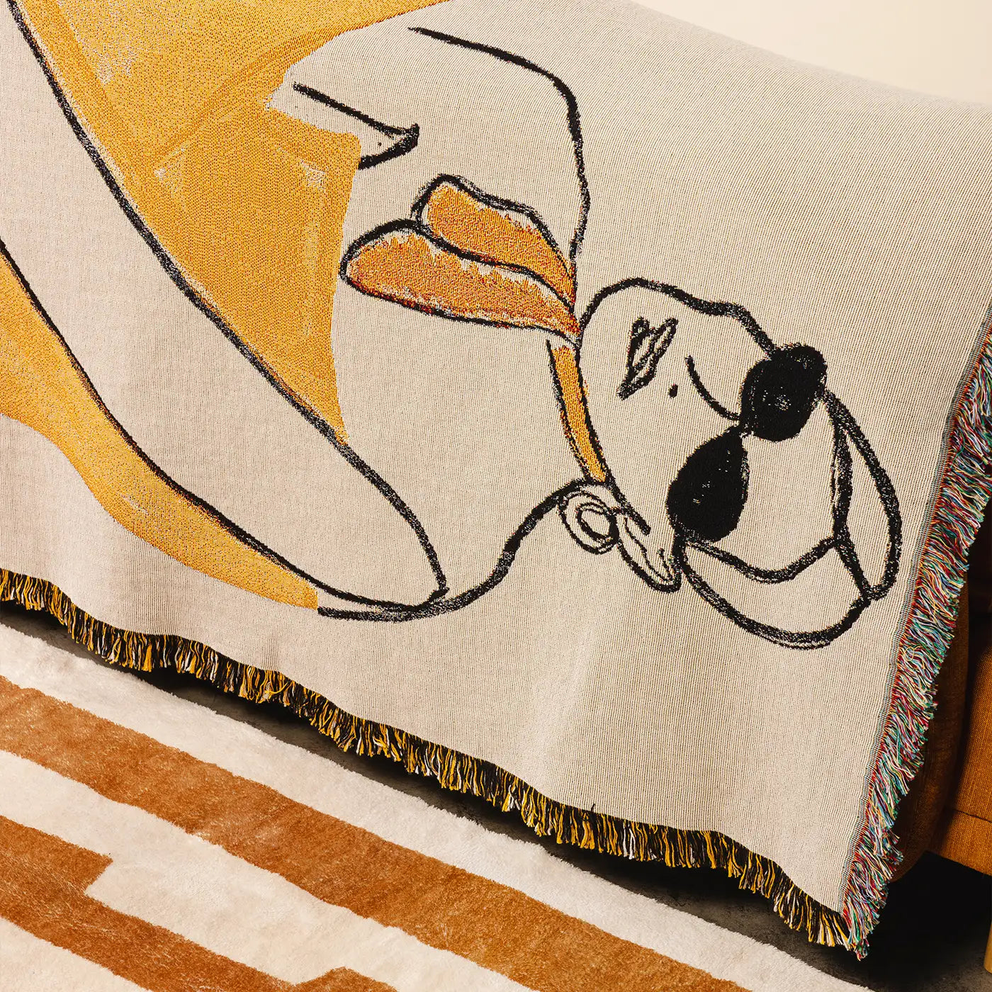 A close-up of a woven throw blanket draped over a chair, featuring a stylized lady design in mustard yellow, black, and cream, with multicolor fringe along the edge.