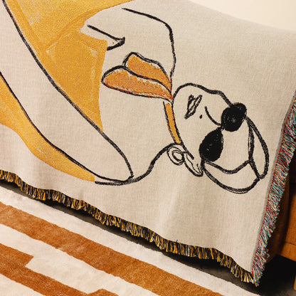 A close-up of a woven throw blanket draped over a chair, featuring a stylized lady design in mustard yellow, black, and cream, with multicolor fringe along the edge.