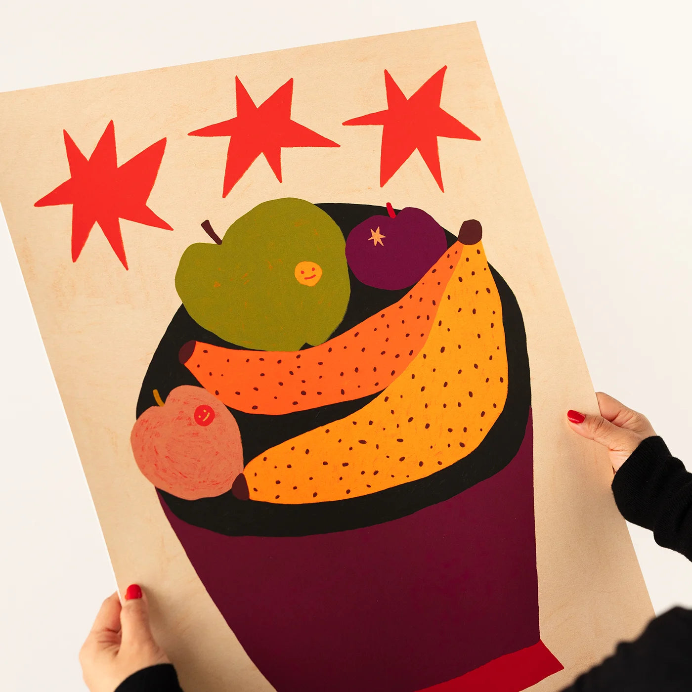 A person holds a large art print featuring a stylized fruit bowl with red stars above, shown against a plain white background.