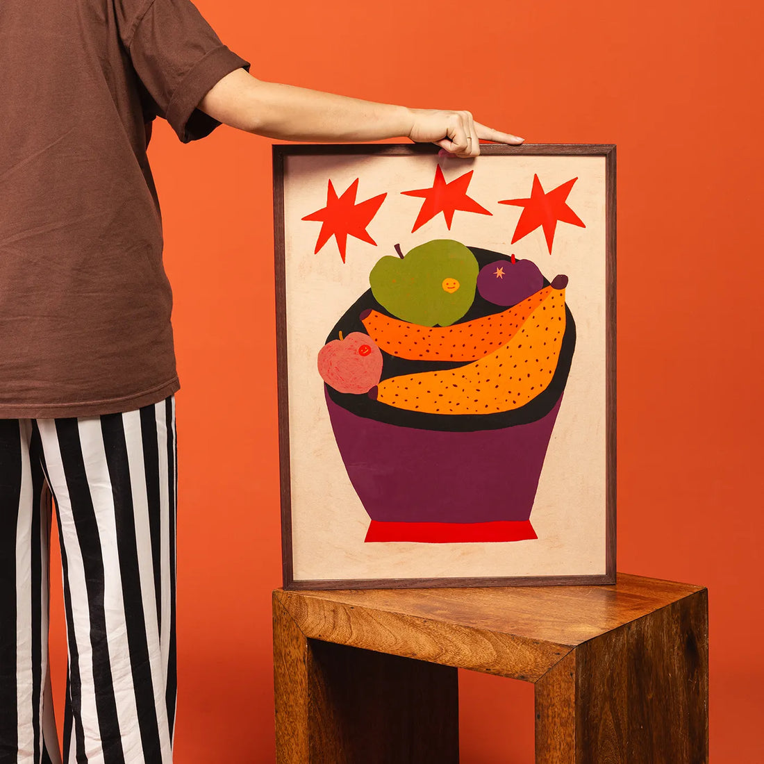 A framed art print featuring a colorful fruit bowl illustration with red stars, held by a person wearing a brown shirt and black-and-white striped pants against a solid orange background.