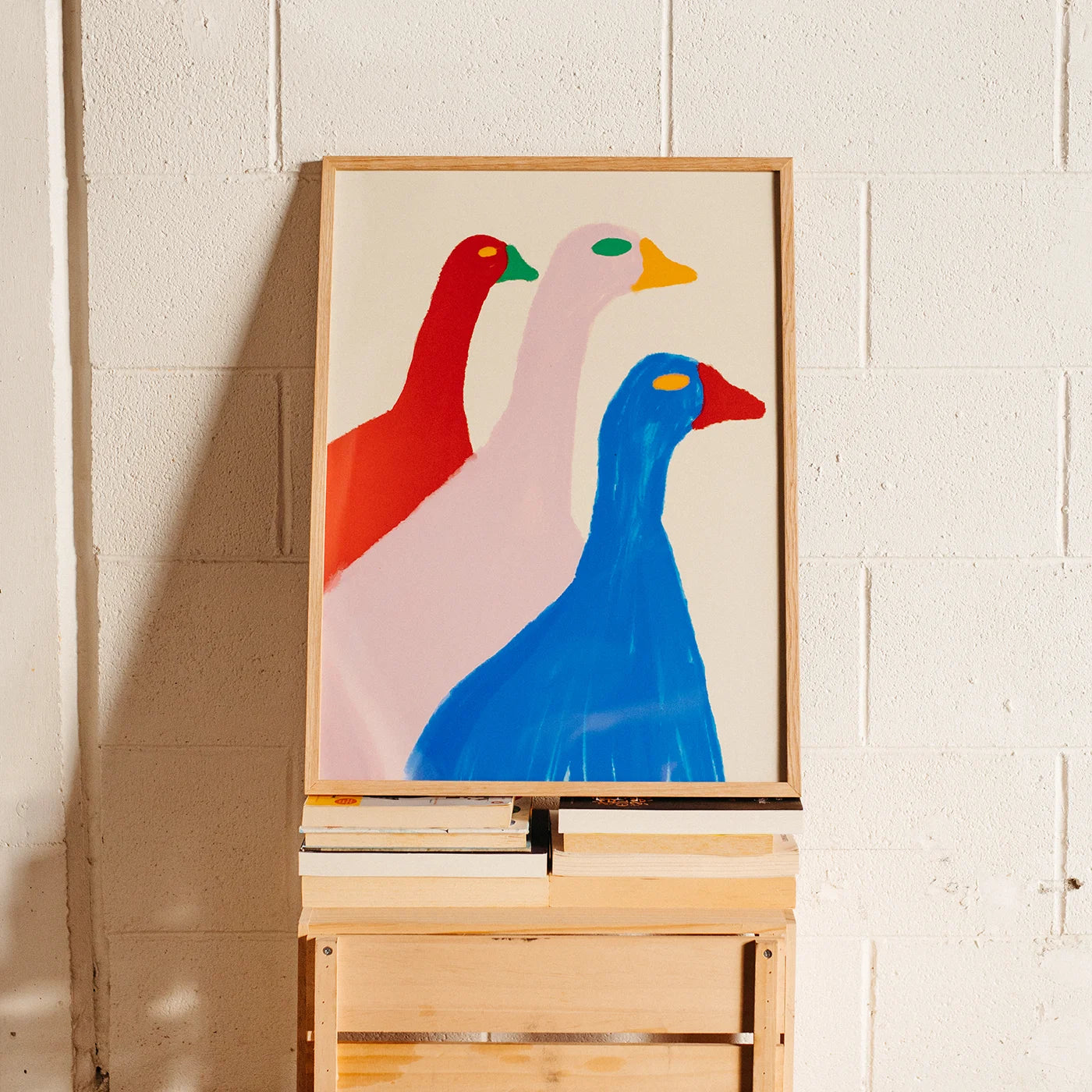 A framed abstract art print depicting three stylized geese in red, pale pink, and blue, displayed on a wooden easel against a white painted brick wall.