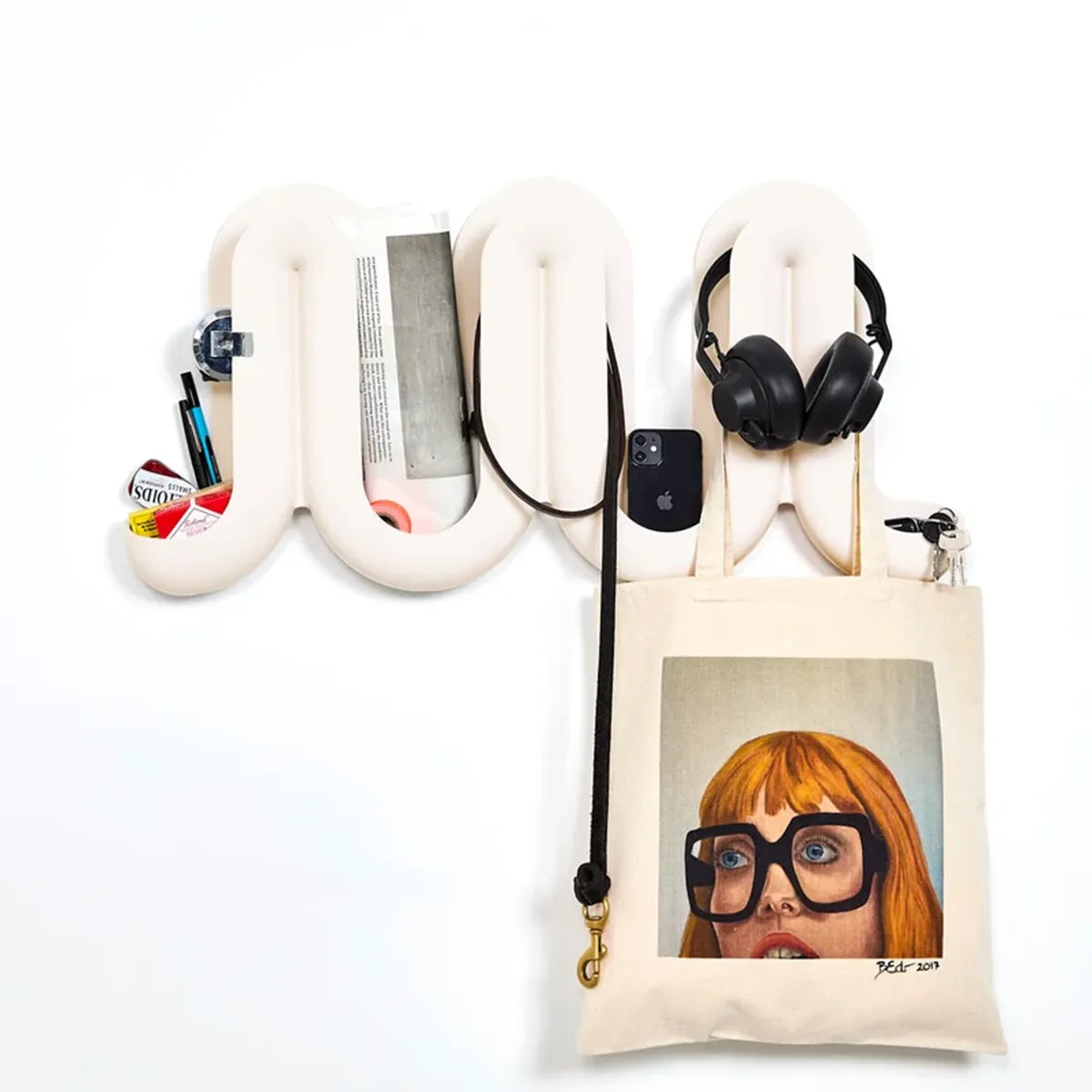A white Heller Swell Wall Catchall, shown holding various items including a tote bag, headphones, a phone, and small accessories. The tote bag features a portrait illustration of a person with orange hair and black glasses.