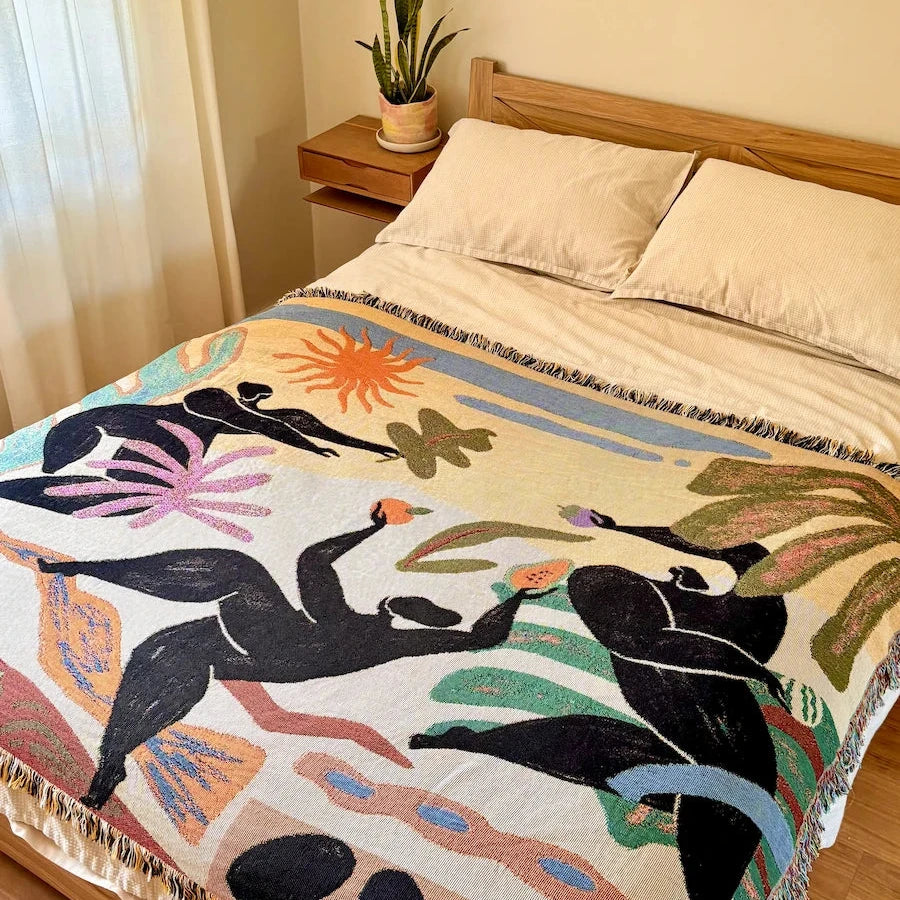 A woven throw blanket bed cover with a colorful abstract bohemian design featuring black figures, orange suns, and green leaves on a cream background, styled on a bed in a warm bedroom setting.