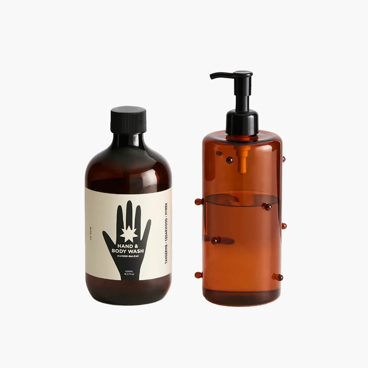 An amber glass hand and body wash set on a light gray background, featuring a dark amber bottle with a black cap and cream label, and a matching translucent amber pump bottle with a black dispenser.