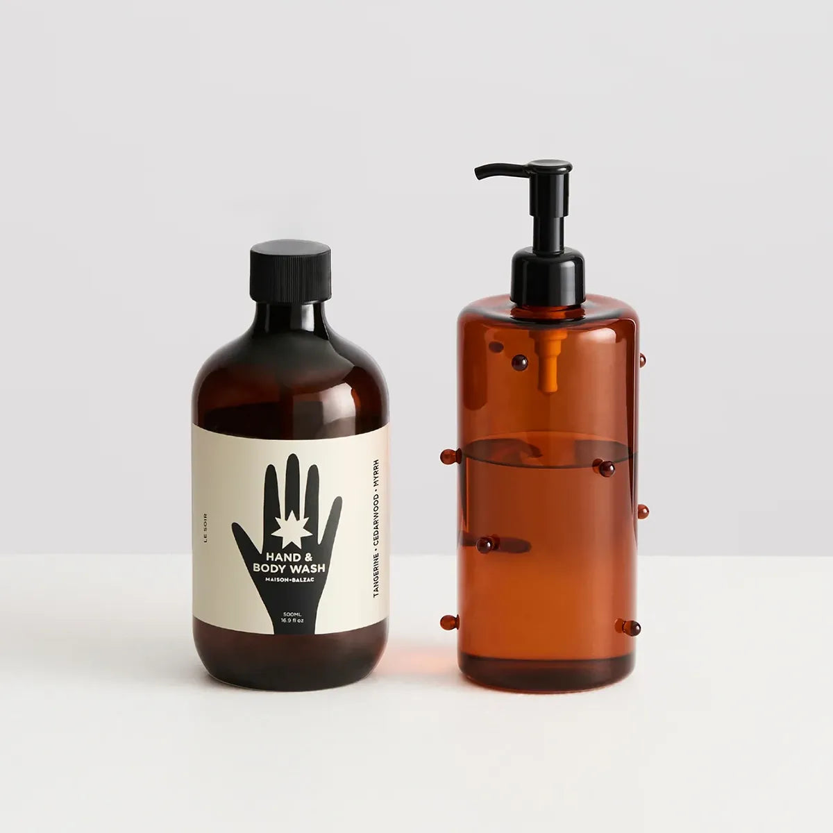 Two amber glass toiletry bottles on a white surface against a light gray background. The left bottle has a black cap and a cream label with a black hand graphic and text reading HAND &amp; BODY WASH.