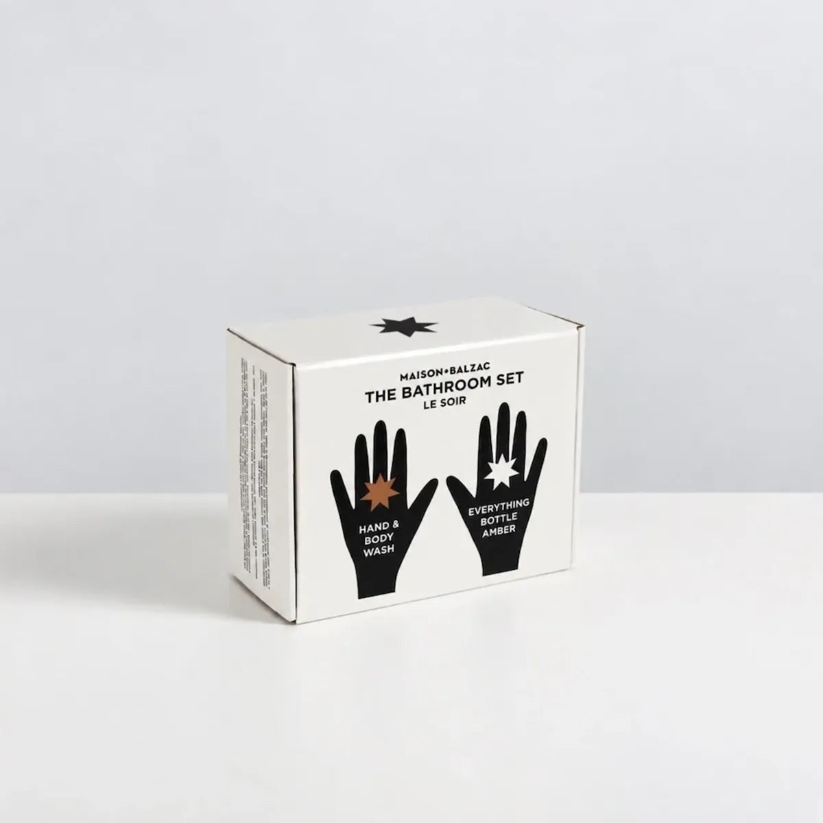 A white cardboard box for Maison &amp; Balzac The Bathroom Set Le Soin, featuring black text and hand graphics on the front, photographed on a clean white surface against a light gray background.