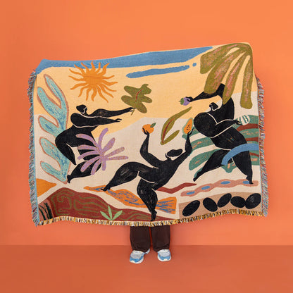 A large woven throw blanket with fringe is held up against a solid orange background. The blanket features a colorful, abstract folk-art style design with black human figures, a yellow sun, and green and blue botanical elements on a cream base.