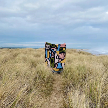 Colorful patterned blanket draped over a grassy dune with ocean and sky in the background