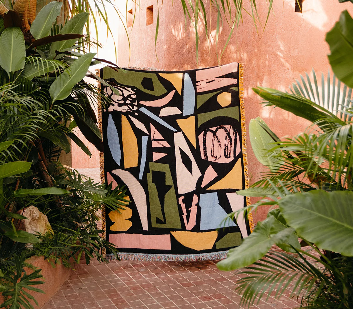 Abstract woven blanket with geometric patterns in front of a pink wall with plants