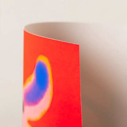 A close-up of an art print corner showing a bright red surface with an abstract blue, purple, and yellow shape, photographed against a soft beige background.