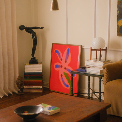 Wall art for living room depicting a framed abstract art print with a bright red background and colorful floral-like shapes leans against a cream paneled wall in a styled living room corner, alongside books, a black figurative sculpture, a side table with a lamp, and a wooden coffee table in the foreground.