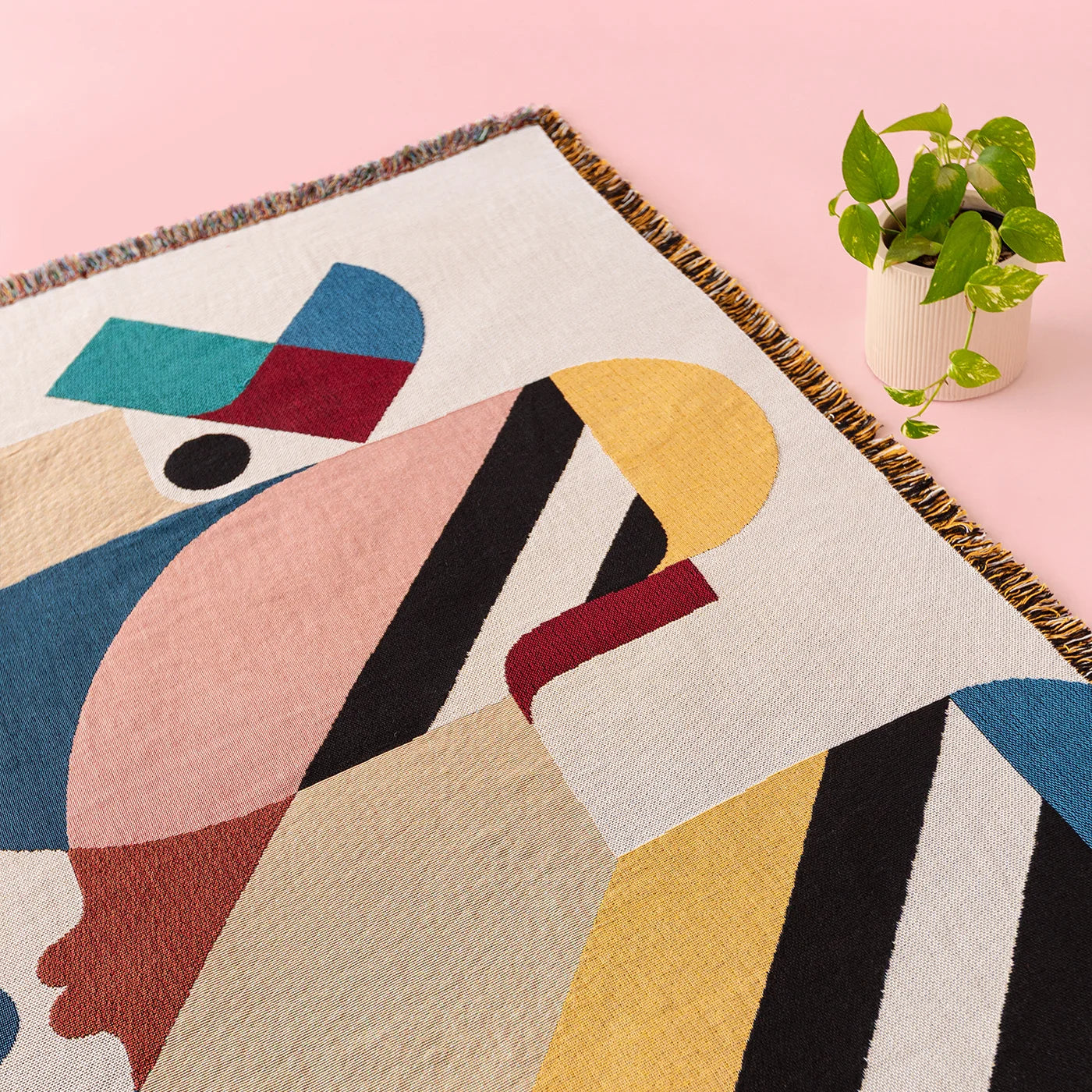 A close-up of a woven throw blanket featuring a colorful abstract geometric design, styled on a light pink surface next to a small potted green plant.