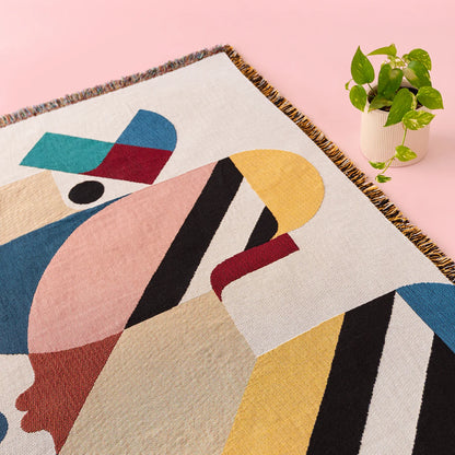 A close-up of a woven throw blanket featuring a colorful abstract geometric design, styled on a light pink surface next to a small potted green plant.