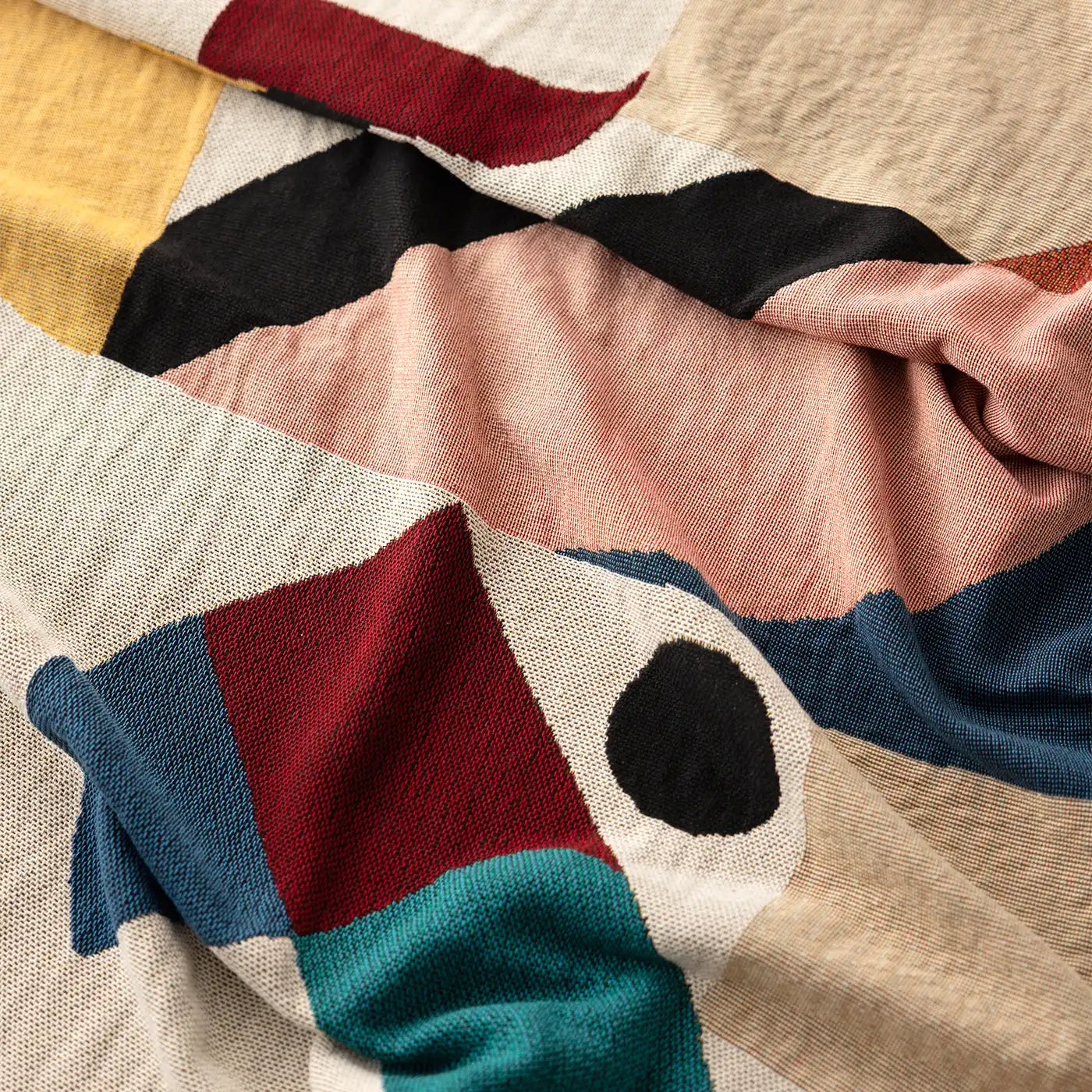 A close-up of the Slowdown Studio Pierre  blanket or throw with an abstract geometric pattern in beige, blush pink, black, blue, teal, and red.