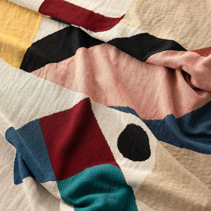 A close-up of the Slowdown Studio Pierre  blanket or throw with an abstract geometric pattern in beige, blush pink, black, blue, teal, and red.